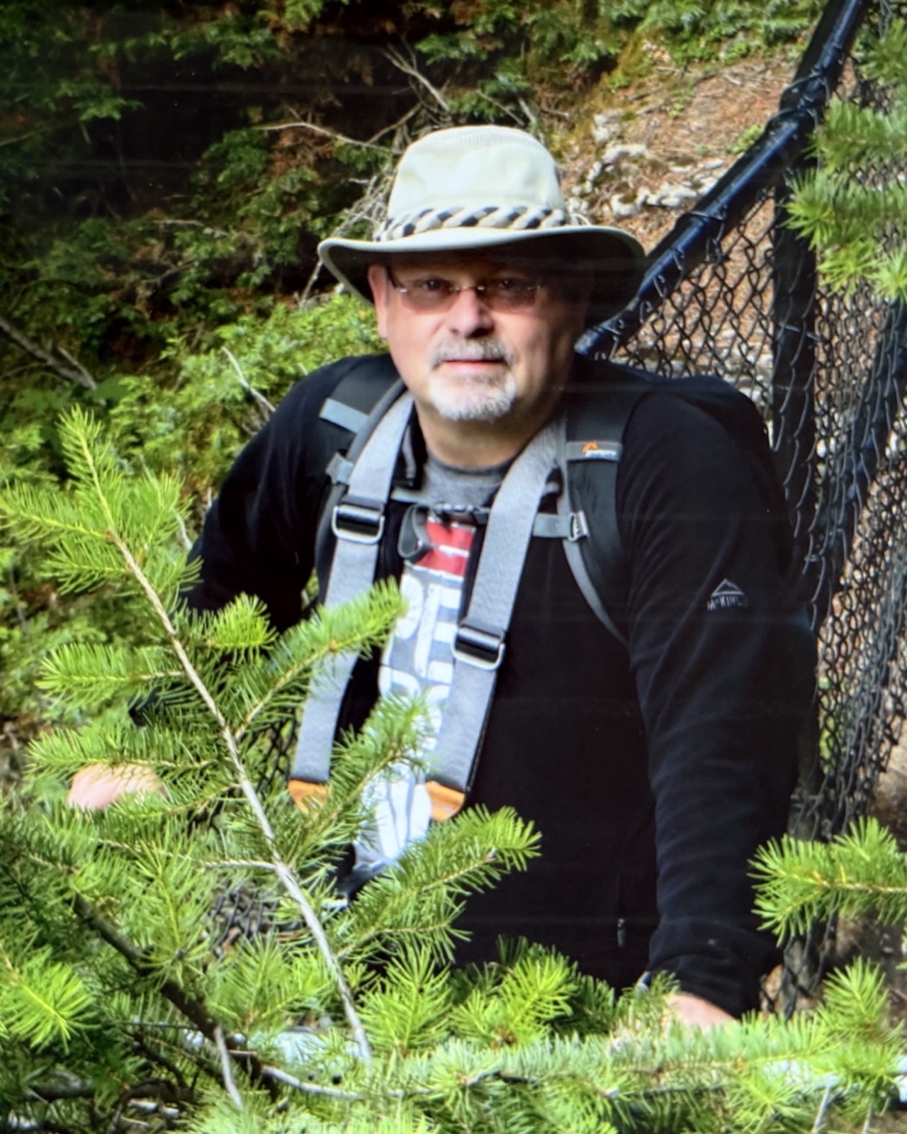 Guy Lahaie coiffé d'un chapeau de randonnée et portant un sac à dos, entre les branches de sapin sur un sentier forestier.