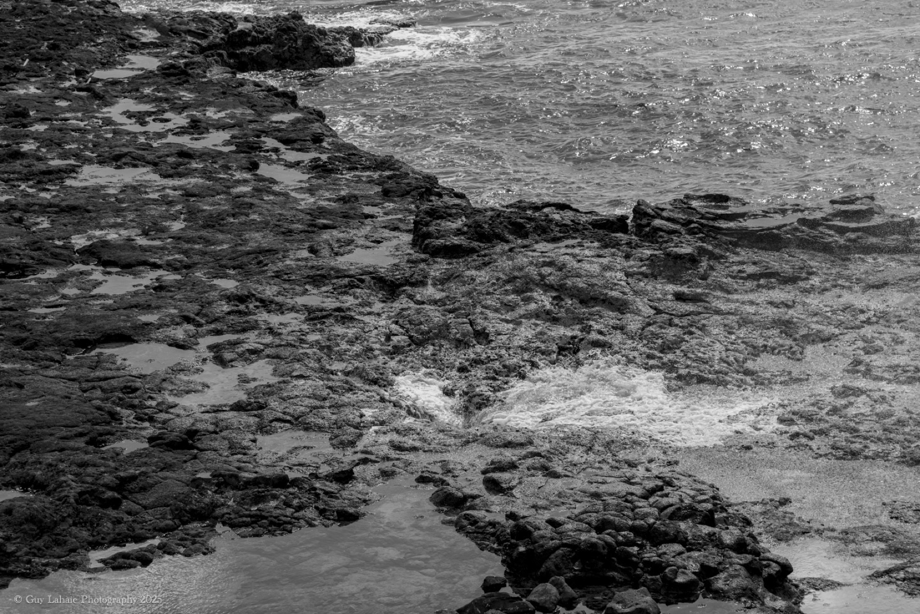 Photo noir et blanc d'une vague se brisant sur une côte rocheuse.